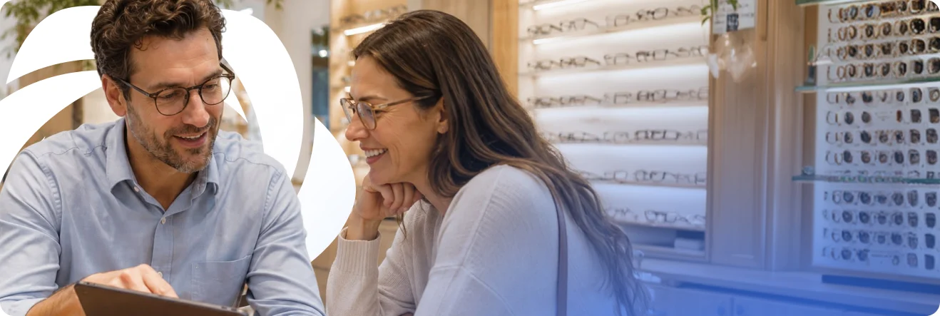 An optician and a customer looking at a tablet together at the counter, in front of a wall of frames.